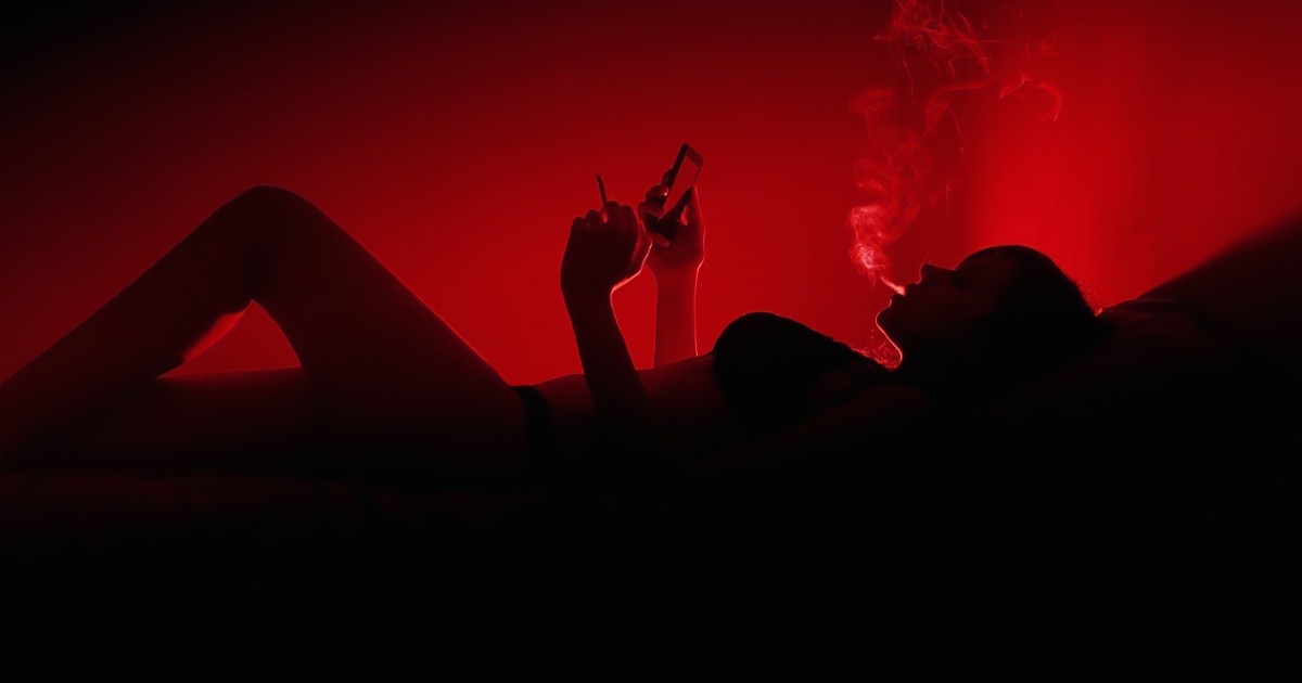 The silhouette of a woman lying on her back shows her exhaling smoke and looking at her cell phone.