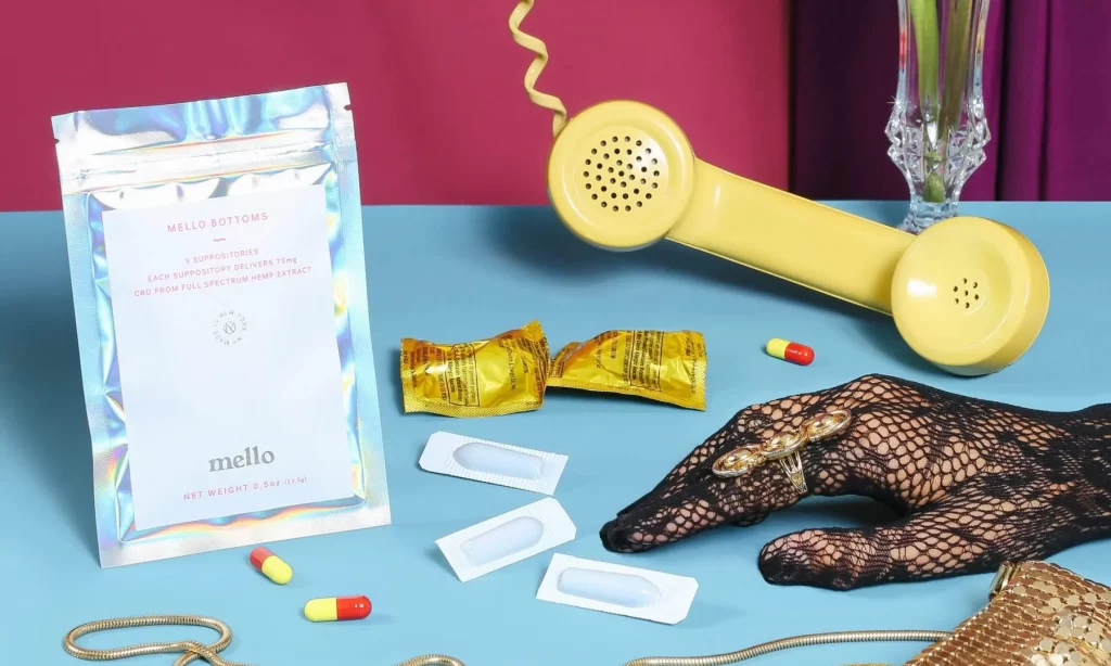 Packaging for Mello Bottoms CBD suppositories on a tabletop along with scattered suppositories, condom wrappers, and an elegant hand near a phone.